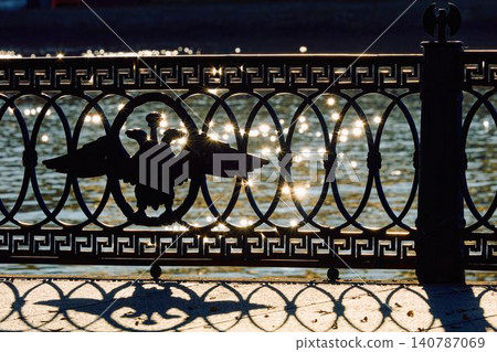 picturesque metal railings with a double-headed eagle and the crown of the Russian Empire on embankment in St. Petersburg, Russia, walking people, sunny autumn day, incredible sun glare Russia 140787069