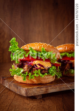 Two burgers with torn beef, tomatoes, fried onions and sauce, laid out on a wooden board, there are no people. 140787220