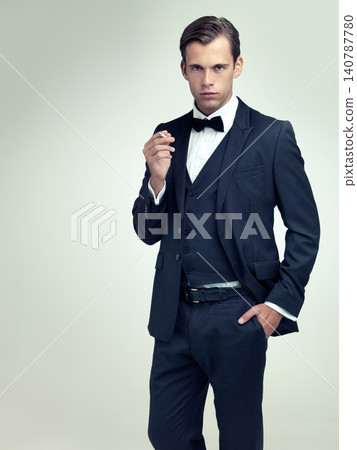 Portrait, serious or young gentleman smoke a cigarette in studio, confidence or vintage fashion by white background. Face, tobacco or man in retro suit or tuxedo, bowtie or hands in pocket for class Portrait, serious or young gentleman smoke a cigarette in studio, confidence or vintage fashion by white background. Face, tobacco or man in retro suit or tuxedo, bowtie or hands in pocket for class 140787780