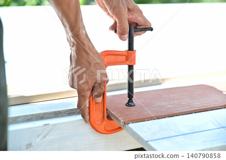 A carpenter's hands tightening a heavy-duty orange metal C-clamp on wood materials during a DIY renovation or construction project. A carpenter's hands tightening a heavy-duty orange metal C-clamp on wood materials during a DIY renovation or construction project. 140790858