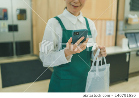 A female supermarket employee in her 50s takes her smartphone out of a transparent bag and looks at it. A female supermarket employee in her 50s takes her smartphone out of a transparent bag and looks at it. 140791650