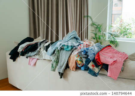 A large, messy pile of colorful baby clothes and garments stacked on a bright sofa in a living room, waiting to be folded or ironed. Stock photo. Housework and laundry concept featuring a big heap of A large, messy pile of colorful baby clothes and garments stacked on a bright sofa in a living room, waiting to be folded or ironed. Stock photo. Housework and laundry concept featuring a big heap of 140794644