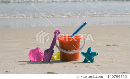 Colorful children's beach sand toys including bucket, shovel, rake and star mold standing on sandy beach with calm sea waves in background. Summer play concept Colorful children's beach sand toys including bucket, shovel, rake and star mold standing on sandy beach with calm sea waves in background. Summer play concept 140794733