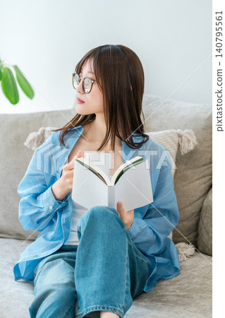 A stylish Asian woman sitting on the living room sofa reading a book, a novel. A stylish Asian woman sitting on the living room sofa reading a book, a novel. 140795561