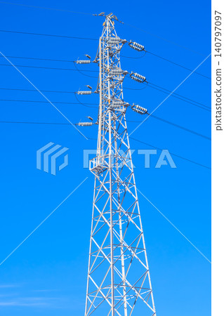 A pure white power transmission tower and numerous power lines stand tall against a vibrant blue sky. 140797097