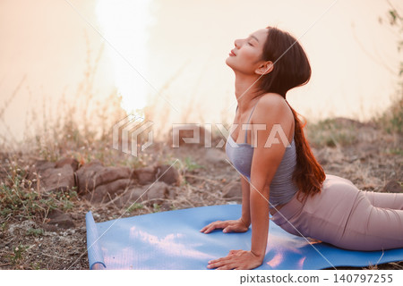 Woman practicing cobra yoga pose outdoors on blue mat near lakeside sunset, focusing on flexibility, spine mobility, mindful breathing, workout recovery, relaxation, wellness, and natural balance. Woman practicing cobra yoga pose outdoors on blue mat near lakeside sunset, focusing on flexibility, spine mobility, mindful breathing, workout recovery, relaxation, wellness, and natural balance. 140797255