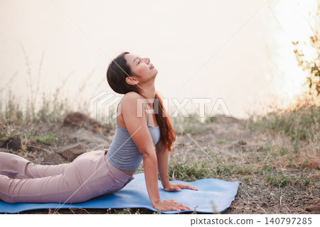 Woman practicing outdoor cobra yoga pose on blue mat near lakeside at sunset, focusing on flexibility, recovery, mindful breathing, back stretch, wellness, relaxation, and healthy lifestyle. Woman practicing outdoor cobra yoga pose on blue mat near lakeside at sunset, focusing on flexibility, recovery, mindful breathing, back stretch, wellness, relaxation, and healthy lifestyle. 140797285