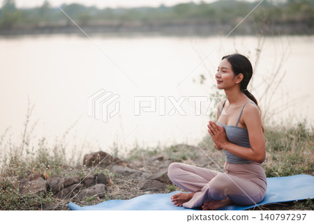 Woman practicing outdoor seated meditation on blue yoga mat by calm lake, hands in prayer, focusing on mindfulness, breathing, nature wellness, balance, relaxation, and peaceful healthy lifestyle. Woman practicing outdoor seated meditation on blue yoga mat by calm lake, hands in prayer, focusing on mindfulness, breathing, nature wellness, balance, relaxation, and peaceful healthy lifestyle. 140797297