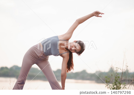 Woman practicing outdoor triangle yoga stretch by lakeside with bright sky, showing flexibility, balance, mobility, mindful breathing, nature wellness, calm focus, and healthy active lifestyle. Woman practicing outdoor triangle yoga stretch by lakeside with bright sky, showing flexibility, balance, mobility, mindful breathing, nature wellness, calm focus, and healthy active lifestyle. 140797298