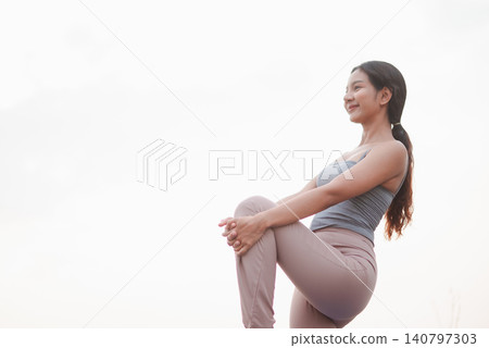 Woman practicing standing knee hug stretch outdoors against bright sky, focusing on balance, mobility, flexibility, warmup, recovery, mindfulness, wellness, and healthy active lifestyle. Woman practicing standing knee hug stretch outdoors against bright sky, focusing on balance, mobility, flexibility, warmup, recovery, mindfulness, wellness, and healthy active lifestyle. 140797303