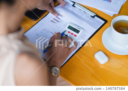 Close-up of hands using calculator on business report with charts beside coffee cup, showing financial analysis, accounting, budget planning, audit data, productivity, and office workflow. Close-up of hands using calculator on business report with charts beside coffee cup, showing financial analysis, accounting, budget planning, audit data, productivity, and office workflow. 140797384