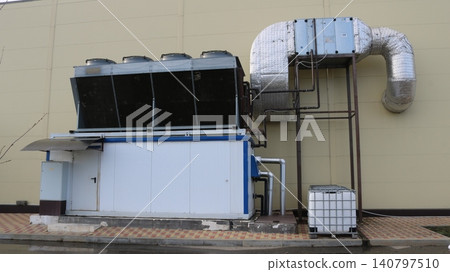 An industrial chiller with a remote condenser is installed against the building wall. The system includes insulated air ducts, cooling fans, and a Eurocube for drainage. The equipment provides climate An industrial chiller with a remote condenser is installed against the building wall. The system includes insulated air ducts, cooling fans, and a Eurocube for drainage. The equipment provides climate 140797510
