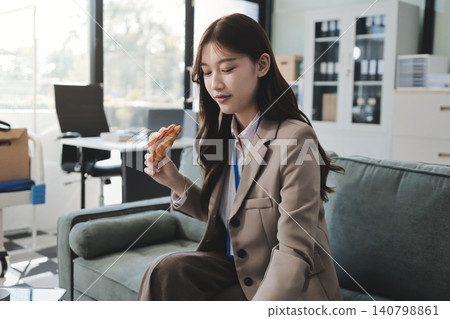 Young asian businesswoman eating a brownie while working from her office on a laptop next to a window with blinds Young asian businesswoman eating a brownie while working from her office on a laptop next to a window with blinds 140798861