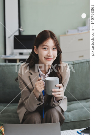 Young asian businesswoman eating a brownie while working from her office on a laptop next to a window with blinds Young asian businesswoman eating a brownie while working from her office on a laptop next to a window with blinds 140798862