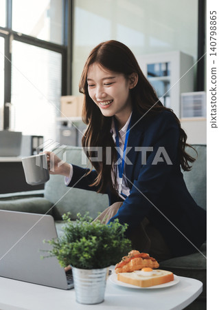 Young asian businesswoman eating a brownie while working from her office on a laptop next to a window with blinds Young asian businesswoman eating a brownie while working from her office on a laptop next to a window with blinds 140798865
