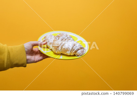 Hand holding plate with fresh croissant sprinkled with powdered sugar on yellow background. Person serving sweet French pastry dessert, minimalist breakfast or bakery concept with copy space Hand holding plate with fresh croissant sprinkled with powdered sugar on yellow background. Person serving sweet French pastry dessert, minimalist breakfast or bakery concept with copy space 140799069