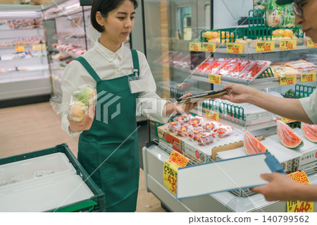 A female staff member in the produce department of a supermarket is sharing information with her colleagues using a tablet. A female staff member in the produce department of a supermarket is sharing information with her colleagues using a tablet. 140799562