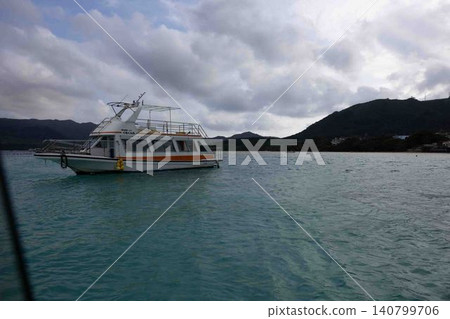A white glass-bottom boat sails across a calm, emerald-green sea, with the cloudy sky encroaching on the landscape. 140799706