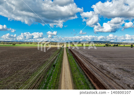 Aerial view of cultivated agricultural fields surrounded by lush green landscape, with a dirt road leading through the farmland, highlighting rural patterns and textures in NSW, Australia. 140800107