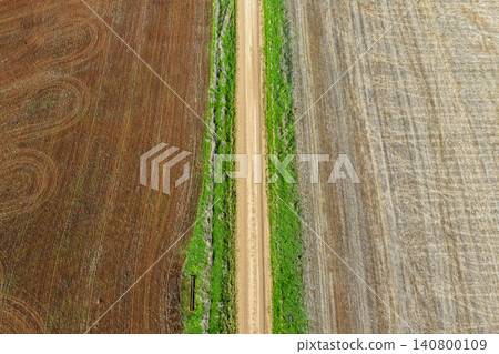 Aerial downward view of a green grass lined dirt road leading through cultivated agricultural fields, showcasing rural farmland patterns and textures beneath soft natural light in NSW, Australia. 140800109
