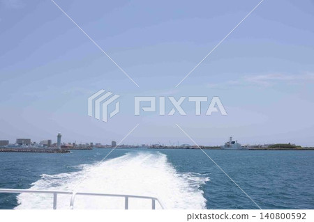 From the ship's deck, one can see the white wake and the blue sea, and in the distance, the harbor town and lighthouse. 140800592