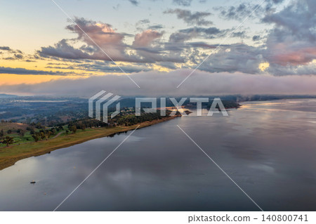 Aerial scene capturing a colourful sunrise over Lake Keepit, with soft dawn light illuminating the shoreline leading to the dam wall as morning fog drifts across the surrounding rural landscape Aerial scene capturing a colourful sunrise over Lake Keepit, with soft dawn light illuminating the shoreline leading to the dam wall as morning fog drifts across the surrounding rural landscape 140800741