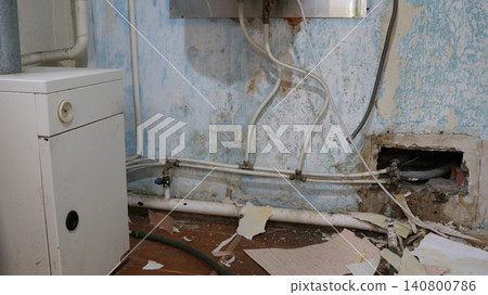 A dilapidated boiler room with a floor-standing boiler and piping against a damp, peeling wall. Construction debris and scraps of old wallpaper litter the floor. A niche in the wall and fittings 140800786