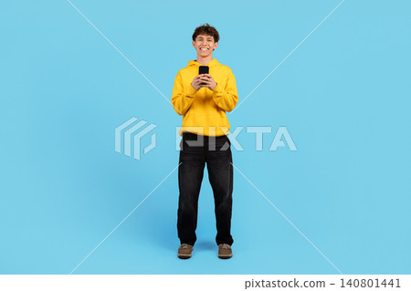 Smiling young guy in yellow hoodie standing with smartphone on blue background Smiling young guy in yellow hoodie standing with smartphone on blue background 140801441