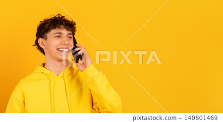 Smiling young guy in hoodie talking on smartphone against yellow studio background Smiling young guy in hoodie talking on smartphone against yellow studio background 140801469