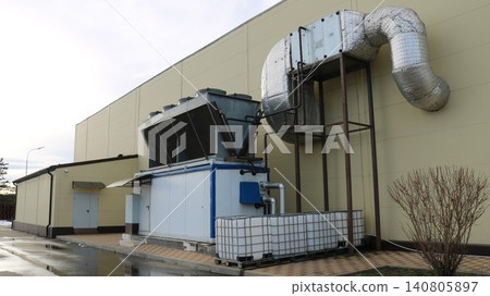 A large-scale industrial air conditioning unit, located outdoors, close to the building's blank wall. In the foreground is the main process station a single-block chiller A large-scale industrial air conditioning unit, located outdoors, close to the building's blank wall. In the foreground is the main process station a single-block chiller 140805897