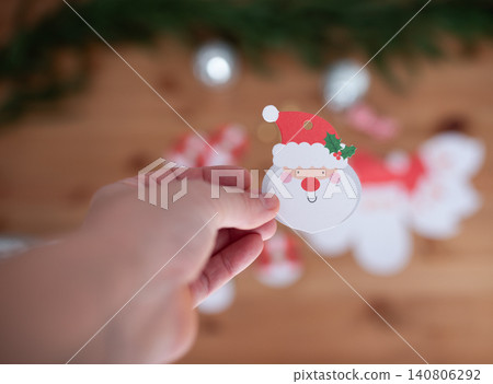 Hand holding a festive Santa Claus paper cutout with holiday decorations in the background, showcasing a cheerful and creative Christmas atmosphere for seasonal celebrations Hand holding a festive Santa Claus paper cutout with holiday decorations in the background, showcasing a cheerful and creative Christmas atmosphere for seasonal celebrations 140806292
