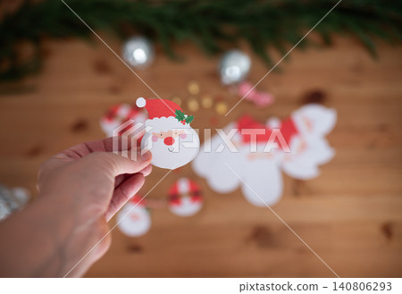 Hand holding a festive Santa Claus paper cutout with red hat, surrounded by colorful decorations and holiday-themed elements on a wooden surface, creating a cheerful atmosphere Hand holding a festive Santa Claus paper cutout with red hat, surrounded by colorful decorations and holiday-themed elements on a wooden surface, creating a cheerful atmosphere 140806293