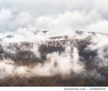 Mist drifts across a brown, heather-covered mountainside under a bright sky. Wisps of white fog obscure the tops of distant, rugged hills. 140806509