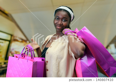 Smiling young African American woman standing in an indoor shopping mall holding bright pink and purple paper bags. She looks confidently at the camera, enjoying a successful day of retail shopping 140807410