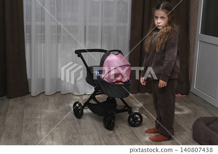 Little girl stands near toy baby stroller in room. Little girl stands near toy baby stroller in room. 140807438
