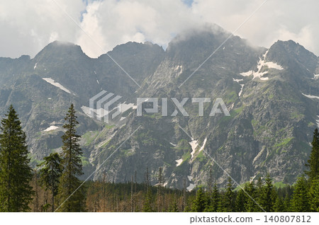 Mountain landscape with rocky peaks and forest in foreground. montane range rising under dramatic clouds. Mountain scenery shows snow patches on slopes, natural summer background, travel concept, Mountain landscape with rocky peaks and forest in foreground. montane range rising under dramatic clouds. Mountain scenery shows snow patches on slopes, natural summer background, travel concept, 140807812