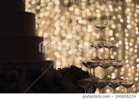 a silhouette shot of small tower of champagne's glass in wedding reception party near by a big cake and golden light bulbs in background. a silhouette shot of small tower of champagne's glass in wedding reception party near by a big cake and golden light bulbs in background. 140808224