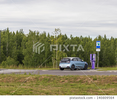 Modern blue compact electric vehicle charging at a public EV station in a green park environment next to a forest. Modern blue compact electric vehicle charging at a public EV station in a green park environment next to a forest. 140809564