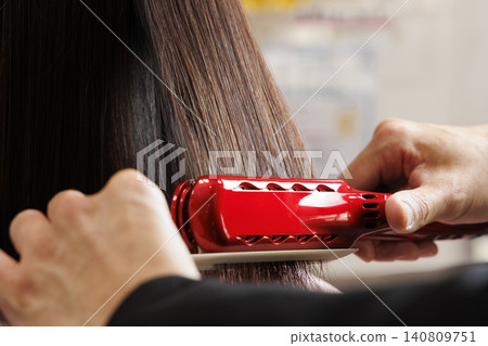 A hairdresser straightens a woman's hair with a red curling iron. A hairdresser straightens a woman's hair with a red curling iron. 140809751