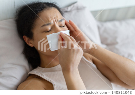 Sick black woman lying in bed and using tissue, blowing her nose Sick black woman lying in bed and using tissue, blowing her nose 140811048