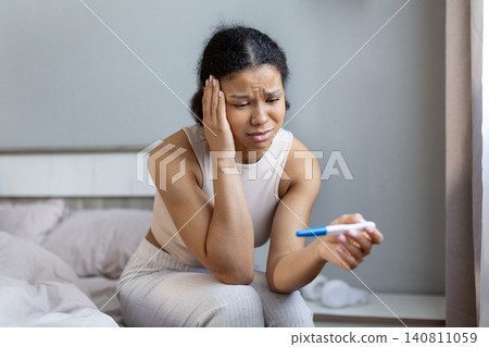 Black woman sitting on bed and looking worried while holding pregnancy test Black woman sitting on bed and looking worried while holding pregnancy test 140811059