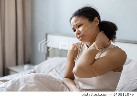 Black woman sitting in bed and holding her neck with concerned expression Black woman sitting in bed and holding her neck with concerned expression 140811079