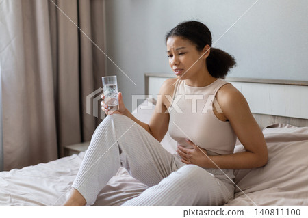 Black woman is sitting on bed holding glass of water and touching her stomach 140811100