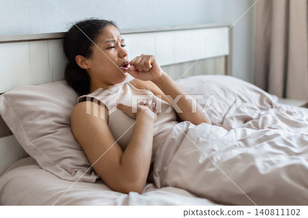 Black woman lying in bed and coughing with one hand on chest Black woman lying in bed and coughing with one hand on chest 140811102