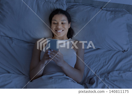 Happy black woman lying in bed at night and using smartphone Happy black woman lying in bed at night and using smartphone 140811175