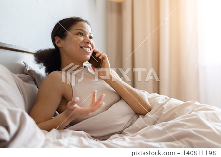 Black woman lying in bed and talking on smartphone, smiling and gesturing Black woman lying in bed and talking on smartphone, smiling and gesturing 140811198