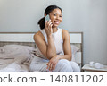 Smiling black woman sitting on bed and talking on smartphone in bedroom 140811202