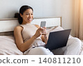 Happy black woman sitting on bed using smartphone beside laptop 140811210