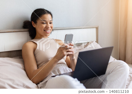 Happy black woman sitting on bed using smartphone beside laptop 140811210