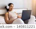 Happy black woman sitting in bed wearing headphones and using laptop 140811221